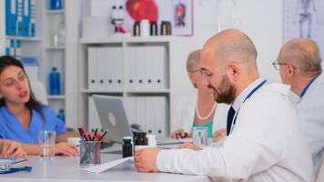 medico controllo elenco di pazienti durante brainstorming, discutere con colleghi seduta a scrivania. squadra di medici parlando di sintomi di malattia nel ospedale ufficio nel sfondo. video