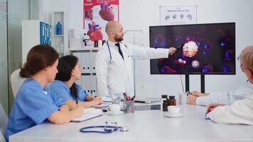 médico presentación virus desarrollo en digital pantalla en frente de colegas señalando en monitor en hospital oficina durante lluvia de ideas. equipo de medicos analizando diagnóstico de pacientes video