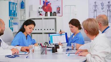 Team of doctors sitting in conference hospital office room discussing about patients list and symptoms of disease, taking notes researching best treatment. Nurse pointing on clipboard brainstorming video