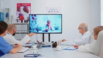 team av doktorer har konferens Sammanträde på skrivbord i sjukhus möte rum diskuterar hälsa frågor med avlägsen specialist kirurg och tar anteckningar. sjuksköterska analyserar diagnos av patienter video