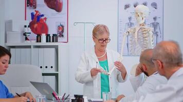 senior kvinna läkare innehav hand skelett modell förklara de arbete av lederna av fingrar på artificiell modell. specialist presenter diagnos till kollegor stående nära skrivbord i sjukhus möte rum video