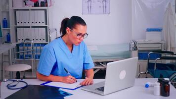 Exausta assistente encarando às computador portátil tela, escrevendo em prancheta, trabalhando a partir de hospital escritório. médico médico dentro remédio uniforme escrevendo Lista do consultado, diagnosticado pacientes, fazer pesquisar. video