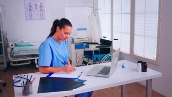 Therapist doctor assistant in uniform writing on clipboard, checking list of patients working on laptop in hospital clinic. Noting consulted patients online, making research, useful information video