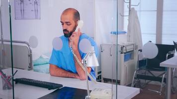 Assistant talking at phone, checking appointment sitting behind a glass wall in hospital office. Medical physician in medicine uniform writing list of consulted, diagnosed patients, making research. video