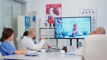 professionnel les collaborateurs en train de regarder pédagogique médical vidéos, Sénior médecin dans blanc manteau séance à bureau dans de face de ordinateur et parlant à collègues par appel, ayant conférence. video