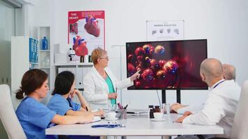 Senior médico apontando em monitor dentro hospital escritório explicando vírus desenvolvimento usando digital tela dentro frente do colegas de trabalho durante debate. equipe do médicos analisando diagnóstico do pacientes video