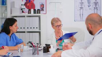 Senior specialist doctor is brainstorming to analyze medical problems pointing on clipboard showing medical statistics. Team of doctors working in hospital office discussing symptoms of disease video