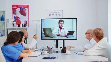 médico personas cuidadosamente escuchando en línea presentación en sala del consejo oficina utilizando moderno tecnología. doctores teniendo conferencia con especialista médico y tomando notas en portapapeles utilizando videollamada. video