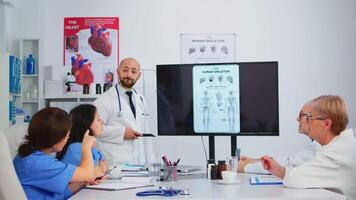 experimentado hombre médico analizando humano esqueleto imagen juntos con calificado colegas en reunión habitación, señalando en monitor. doctores que se discute diagnóstico acerca de tratamiento de pacientes video