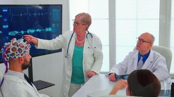 cientista fazendo cérebro análise usando fone de ouvido com sensores em médico dentro hospital encontro sala. monitor mostra moderno cérebro estude enquanto equipe do cientista ajusta a dispositivo trabalhando dentro conferência escritório video