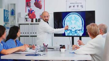 experimentado hombre médico analizando el resultado de digital radiografía imagen juntos con calificado colegas en reunión habitación, señalando en monitor. doctores que se discute diagnóstico acerca de tratamiento de pacientes video