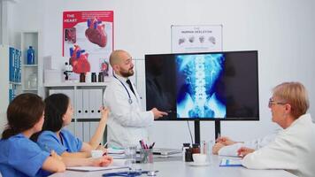 calificado cabeza médico en blanco Saco sentado en hospital sala del consejo explicando girar funciones señalando en digital radiografía imagen. que se discute síntomas de enfermedad para más lejos tratamiento video