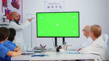 Arzt erklären Treffen Themen durch Vorschau auf ein Grün Bildschirm Monitor im Krankenhaus Konferenz Zimmer. Mannschaft von Ärzte mit vielfältig Alter Brainstorming suchen beim Attrappe, Lehrmodell, Simulation Anzeige mit Grün Chroma Taste. video