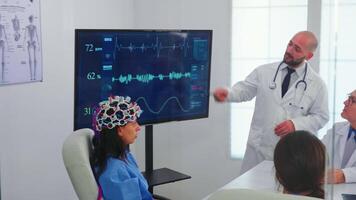 sjuksköterska bär headsetet med sensorer under neurovetenskap experimentera i medicinsk konferens team. övervaka visar modern hjärna studie medan team av forskare justerar de enhet arbetssätt i sjukhus styrelserum video