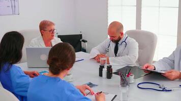 Mature doctor presenting health report during meeting with coworkes in hospital conference room. Clinic expert therapist talking with colleagues about disease, medicine professional video