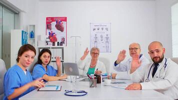 grupo do especialista médicos tendo reunião, discutindo em Webcam sentado às escrivaninha dentro médico conferência hospital quarto usando moderno tecnologia. profissional equipe olhando às Câmera e falando video