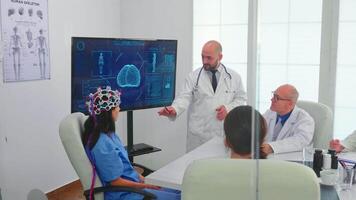 fêmea enfermeira vestindo escaneando fone de ouvido para cérebro atividade durante experimentar e médico dizendo diagnóstico. monitor mostra moderno cérebro estude enquanto equipe do cientista ajusta a dispositivo dentro hospital escritório video