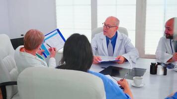 Mature woman doctor pointing on clipboard during briefing with coworkers working in hospital meeting room. Clinic expert therapist talking with colleagues about disease, medicine professional video