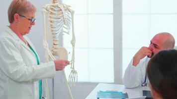 medico medico donna insegnamento anatomia utilizzando umano scheletro modello in piedi nel ospedale conferenza ufficio. clinica esperto terapista parlando con colleghi di patologia, medicina professionale. video