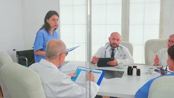 Woman medical assistant using clipboard in hospital conference room with coworkers explaining disease development. Clinic expert therapist talking with colleagues about disease, medicine professional video