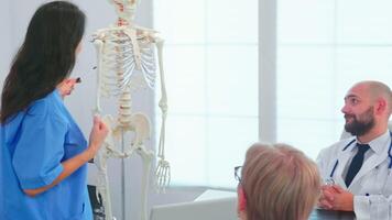 femmina infermiera dimostrando su scheletro nel davanti di medico chirurghi nel conferenza camera. clinica esperto terapista parlando con colleghi di patologia, medicina professionale video