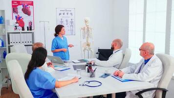 Young female assistent pointing on skeleton in front of medical surgeons in conference room. Clinic expert therapist talking with colleagues about disease, medicine professional video