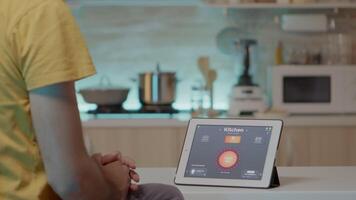 Mens op zoek Bij tablet met intelligent software geplaatst Aan keuken tafel controlerend licht met hoog tech sollicitatie. persoon gebruik makend van kladblok met slim huis app in huis automatisering systeem video