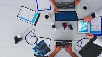 topo Visão do especialista médico analisando humano cérebro usando tábua em flatlay sentado em escrivaninha dentro clínica com médico equipamento. assistentes levando notas em computador portátil e prancheta, pesquisando em cópia de espaço video