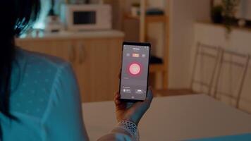 femmina Tenere smartphone con illuminazione controllo applicazione, svolta su il luci seduta nel cucina Casa con automazione illuminazione sistema. donna utilizzando alto tecnologia Software, senza fili interruttore su bulbi video