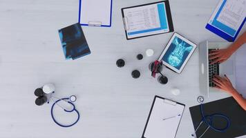 Top view of doctor closing laptop and analysing digital x-ray using tablet, working in medical office sitting on desk with stethoscope and digital devices. Studying on clinic on copy space, flat lay video