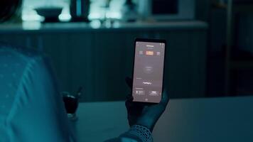 kvinna Sammanträde i hus med automatisering ljus systemet innehav smartphone vändning på ljus använder sig av röst aktiverad Ansökan. person kontrollerande rum atmosfär och energi effektivitet med smart Hem app video