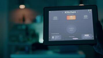 homme en portant tablette avec éclairage contrôle application, tournant sur le lumières séance dans cuisine maison avec automatisation éclairage système. la personne en utilisant haute La technologie Logiciel travail éloigné à portable video