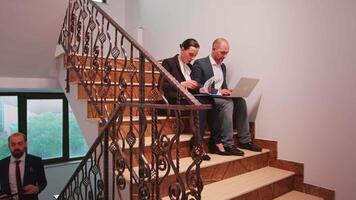 Office colleagues helping coworking at difficult deadline finance project sitting on stairs in corporate company building typing on laptop. Group of businesspeople walking in financial workplace. video