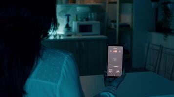 persona guardare a mobile Telefono con illuminazione controllo App seduta nel cucina di Casa con automazione leggero sistema, svolta su lampadina utilizzando voce comando. donna monitoraggio elettricità efficienza video