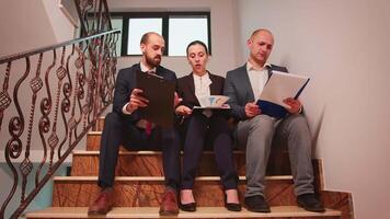 Project team of business workers looking on documents sitting on stairs explaining finance project doing overtime. Group of company manager and office executive on busy stairs of workplace. video