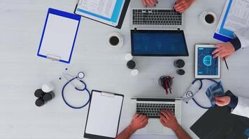 Top view of doctor explaining to resident physician patient problems using tablet and x-ray sitting at desk with stethoscope and digital device all around on copy space, flat lay. video