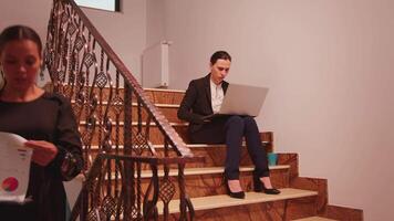 Employee using laptop overworking on deadline sitting on stairs in finance corporate company while colleagues leaving office workplace. Professional businesspeople working in modern financial building video