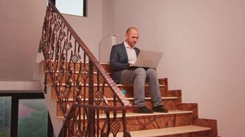 Exhausted businessman using laptop working alone on difficult deadline sitting on stair overtime. Executive manager doing overtime at corporate job on staircase while colleagues leaving office building video