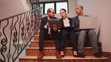 Group of business partners making plans of work sitting on staircase explaining finance project using laptop and tablet analysing reports. Team of company manager and office executive on busy stairs. video