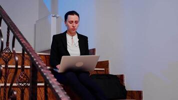 Overworked tired businesswoman with headache working on difficult project deadline using laptop sitting on staircase of business building. Serious entrepreneur being on corporate job late at night. video