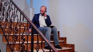 Overworked tired businessman reading project deadline during phone call with corporate manager. Serious entrepreneur working on job sitting on staircase of business building late at night. video