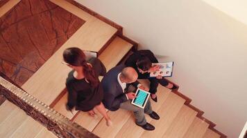 Top view of project team working on stairs sitting down using tablet while colleagues moving up. Colleague entrepreneur working together in evening at corporate job explaining difficult project. video