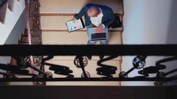 Top view of office executive checking reports from clipboard using laptop sitting on stairs in corporate building working ovretime. Businesspeople doing overtime making plans for deadline project. video