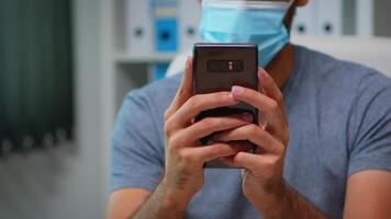 Close up of man with face mask typing text messages on smartphone sitting in workplace during coronavirus. Freelancer working in new normal office chatting talking writing using internet technology video