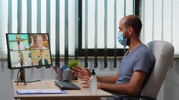 Man with face mask in office talking on call with remote team during coronavirus epidemic. Freelancer discussing chatting having virtual online conference, meeting, using internet technology video