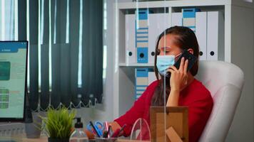 Frau reden auf Telefon tragen medizinisch Gesicht Maske im das Büro respektieren Sozial Distanzierung. Freiberufler Arbeiten im Neu normal Arbeitsplatz chatten mit entfernt Mannschaft Sprechen auf Smartphone video