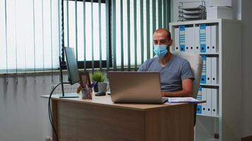 Man with mask having online meeting conference in modern office using laptop. Freelancer working in new normal office workplace chatting talking during virtual webinar, using internet technology video