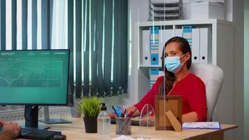 Woman with protection mask working and looking at camera sitting in front of pc. Employees in workspace in corporate company typing on computer keyboard looking at desktop respecting social distancing video