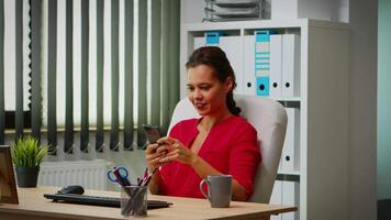 Hispanic lady chatting using smartphone in business office. Entrepreneur working in professional workspace, workplace in personal corporate company typing on mobile phone in front of computer video