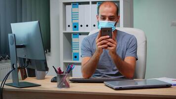 Office worker with protective mask using phone typing sitting on modern office during coronavirus. Freelancer working in new normal workplace chatting talking writing using mobile internet technology video